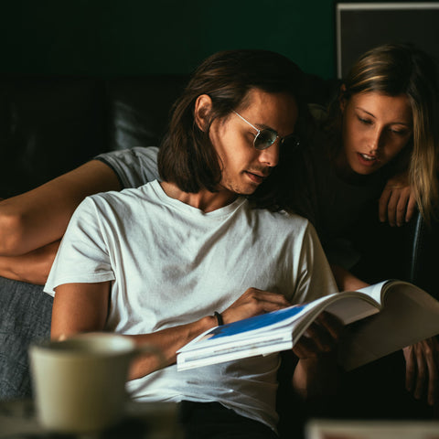 Two people sitting together, one reading a book and the other looking on, in a dimly lit room.