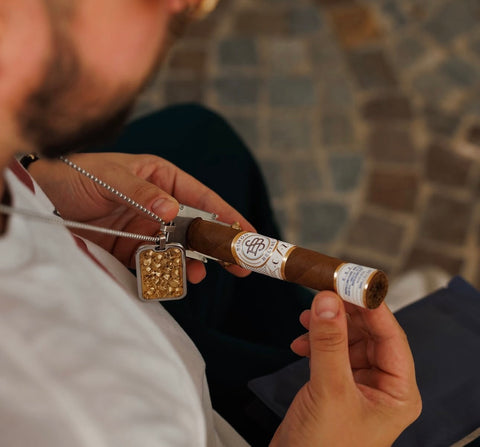 Person holding a cigar with a decorative chain and gold pendant.