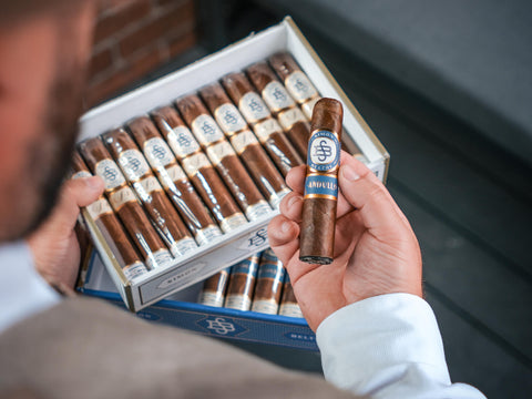 Person holding a cigar with a box of cigars in the background