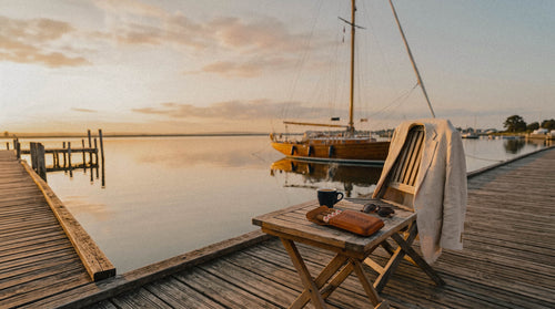 A wooden folding chair on a dock by the water at sunset, with a small table holding two dark mugs and a brown leather Toro Puro accessory case, and a linen jacket draped over the chair. A sailboat is moored in the calm water in the background.